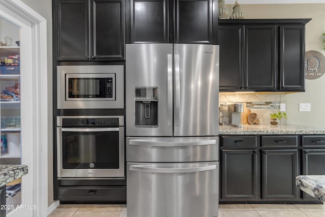 kitchen featuring stainless steel appliances, backsplash, dark cabinetry, light stone countertops, and light tile patterned flooring