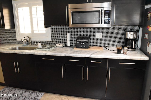 kitchen with a sink, dark cabinetry, backsplash, light stone countertops, and stainless steel microwave