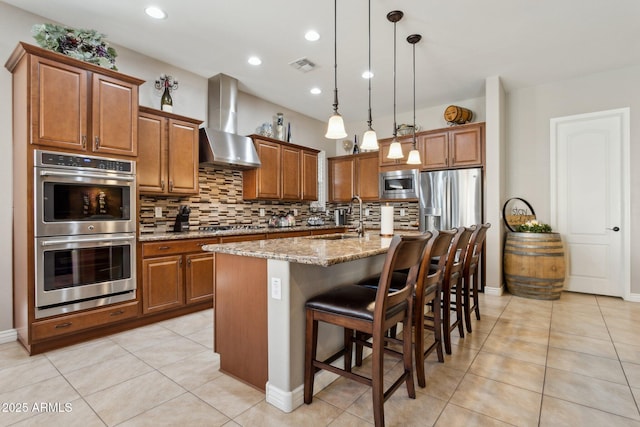 kitchen featuring pendant lighting, wall chimney range hood, a kitchen island with sink, appliances with stainless steel finishes, and sink