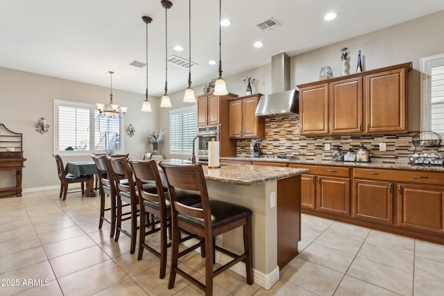 kitchen featuring decorative light fixtures, wall chimney exhaust hood, a kitchen bar, light stone countertops, and a kitchen island with sink