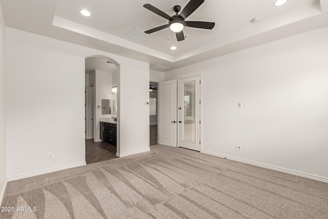 unfurnished bedroom featuring a tray ceiling, carpet flooring, arched walkways, recessed lighting, and baseboards