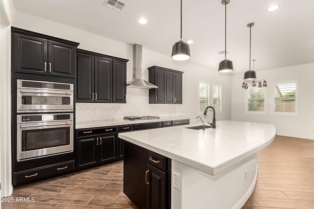 kitchen featuring a sink, wall chimney range hood, wood finish floors, dark cabinets, and healthy amount of natural light