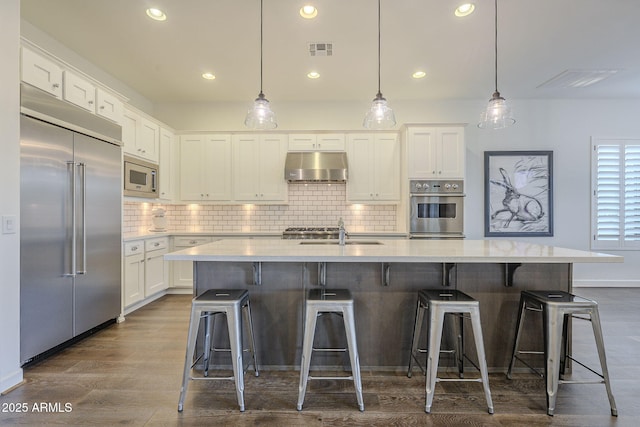 kitchen with a center island with sink, white cabinetry, built in appliances, and decorative light fixtures