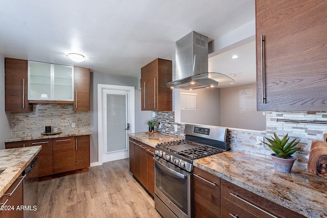 kitchen with island range hood, decorative backsplash, light wood-style flooring, glass insert cabinets, and appliances with stainless steel finishes