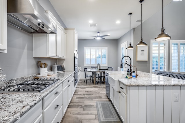 kitchen with wall chimney range hood, hanging light fixtures, a center island with sink, white cabinetry, and light hardwood / wood-style floors