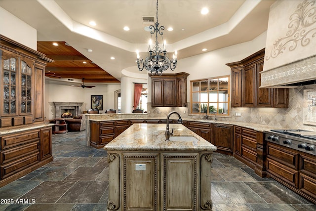 kitchen featuring sink, a kitchen island with sink, light stone counters, a tray ceiling, and stainless steel gas stovetop