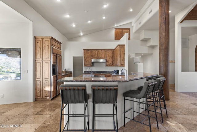 kitchen featuring under cabinet range hood, a peninsula, a breakfast bar, brown cabinets, and dark stone counters