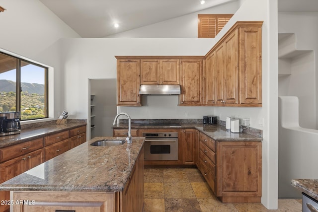 kitchen featuring oven, stone tile floors, under cabinet range hood, a sink, and dark stone counters