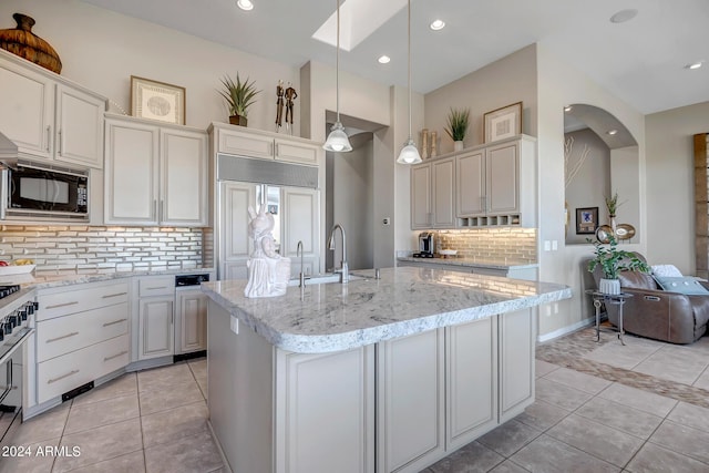 kitchen with sink, built in appliances, hanging light fixtures, an island with sink, and light stone countertops