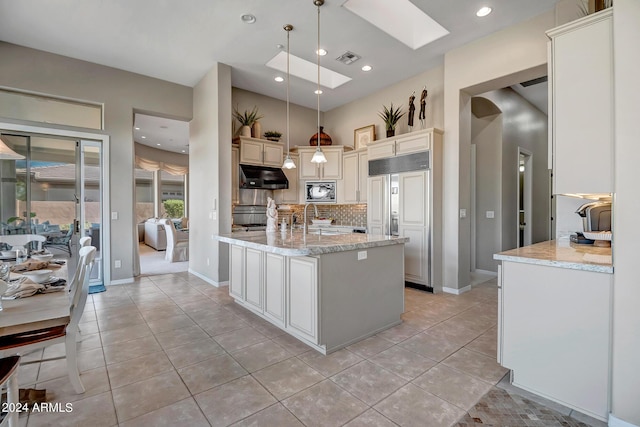 kitchen featuring a skylight, decorative light fixtures, an island with sink, built in appliances, and light stone counters