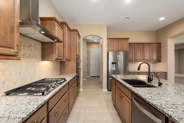 kitchen featuring arched walkways, wall chimney exhaust hood, light stone counters, appliances with stainless steel finishes, and a sink