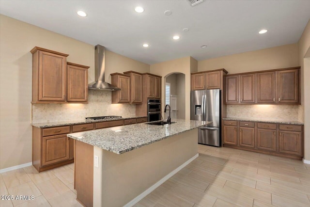 kitchen with a center island with sink, light stone counters, brown cabinets, stainless steel appliances, and wall chimney range hood