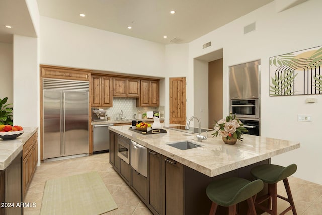 kitchen with sink, tasteful backsplash, a center island with sink, light tile patterned floors, and appliances with stainless steel finishes