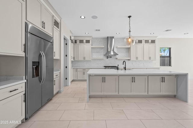 kitchen featuring built in refrigerator, a kitchen island with sink, open shelves, wall chimney exhaust hood, and light countertops