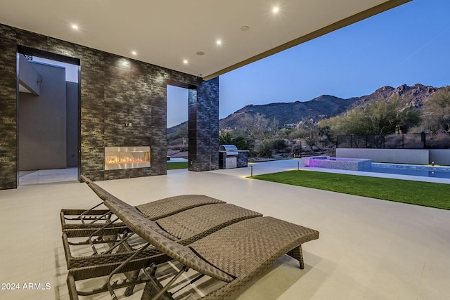 patio terrace at dusk with an outdoor stone fireplace, area for grilling, a fenced in pool, and a mountain view