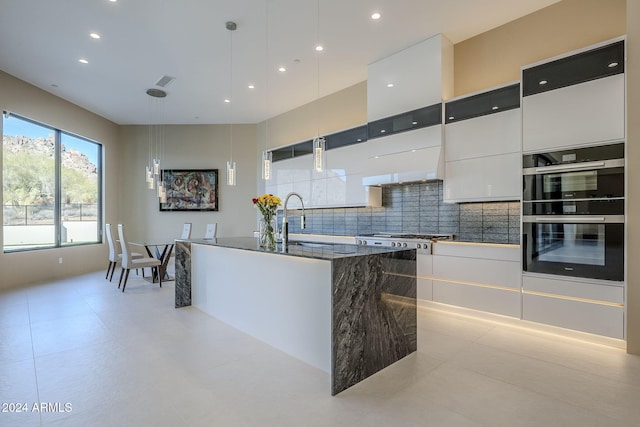 kitchen featuring backsplash, white cabinets, a center island with sink, double oven, and decorative light fixtures