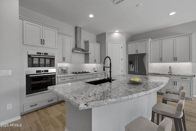 kitchen with appliances with stainless steel finishes, sink, a kitchen island with sink, light stone counters, and wall chimney range hood