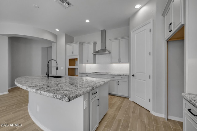 kitchen with light stone counters, sink, wall chimney exhaust hood, and a center island with sink