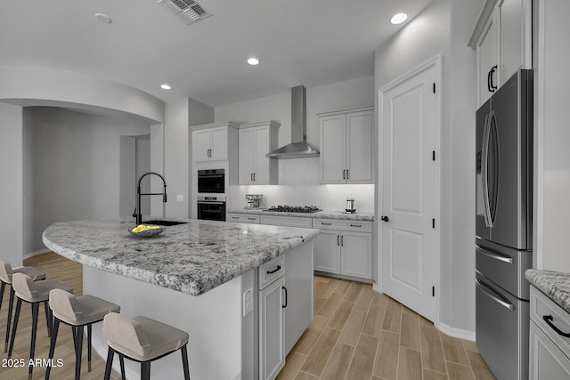 kitchen with stainless steel refrigerator, sink, a kitchen island with sink, light stone counters, and wall chimney range hood