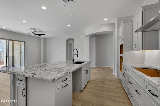 kitchen with sink, white cabinetry, an island with sink, light stone countertops, and wall chimney range hood