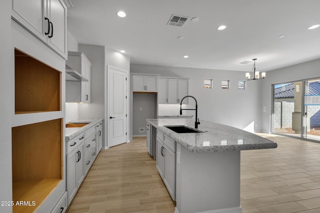 kitchen featuring an island with sink, sink, hanging light fixtures, light stone countertops, and an inviting chandelier