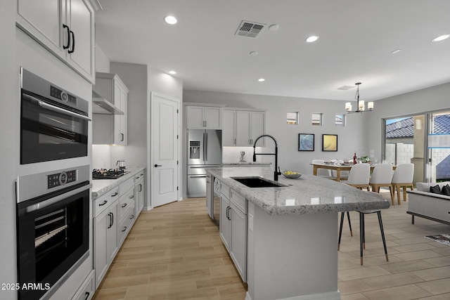 kitchen with sink, appliances with stainless steel finishes, hanging light fixtures, light stone countertops, and an island with sink