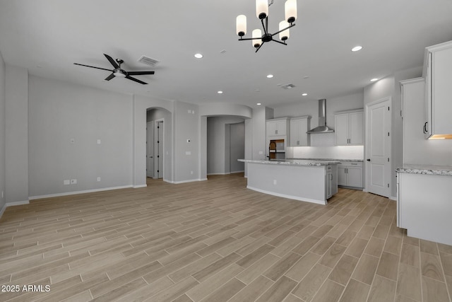 kitchen with light hardwood / wood-style flooring, a kitchen island with sink, wall chimney range hood, ceiling fan with notable chandelier, and white cabinets