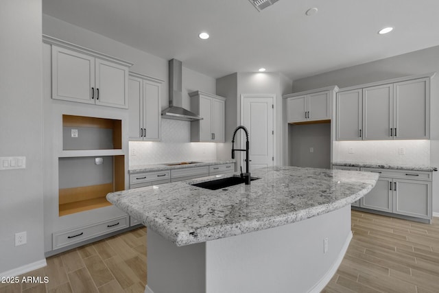 kitchen with wall chimney range hood, sink, light stone counters, black electric stovetop, and an island with sink