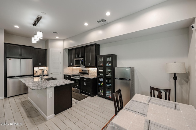 kitchen with sink, appliances with stainless steel finishes, a kitchen island with sink, light stone counters, and tasteful backsplash