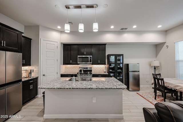 kitchen featuring pendant lighting, sink, a kitchen island with sink, stainless steel appliances, and light stone countertops