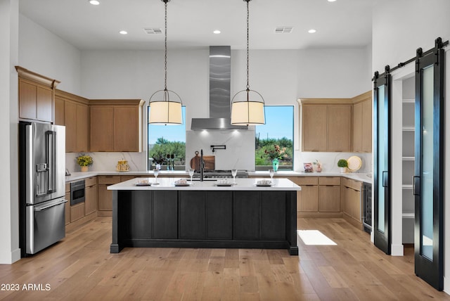 kitchen featuring tasteful backsplash, an island with sink, wall chimney range hood, a barn door, and high end refrigerator