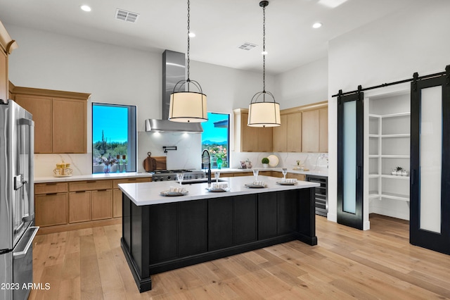 kitchen featuring pendant lighting, wall chimney range hood, stainless steel fridge, a barn door, and a kitchen island with sink