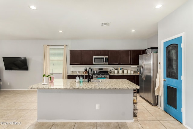 kitchen featuring dark brown cabinetry, light stone counters, a center island with sink, light tile patterned floors, and appliances with stainless steel finishes