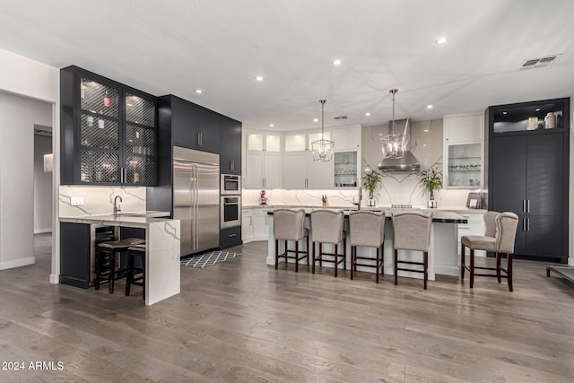 kitchen with wall chimney exhaust hood, a large island with sink, pendant lighting, a kitchen bar, and appliances with stainless steel finishes