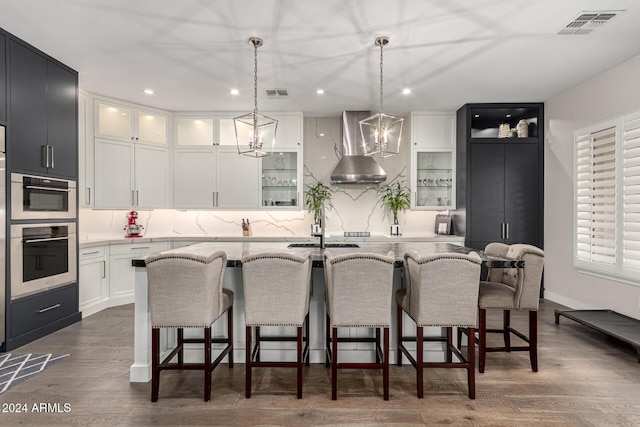 kitchen featuring decorative light fixtures, wall chimney range hood, an island with sink, and dark wood-type flooring