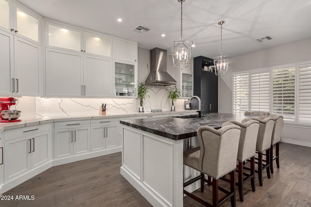 kitchen featuring dark wood-type flooring, wall chimney range hood, dark stone countertops, an island with sink, and white cabinets