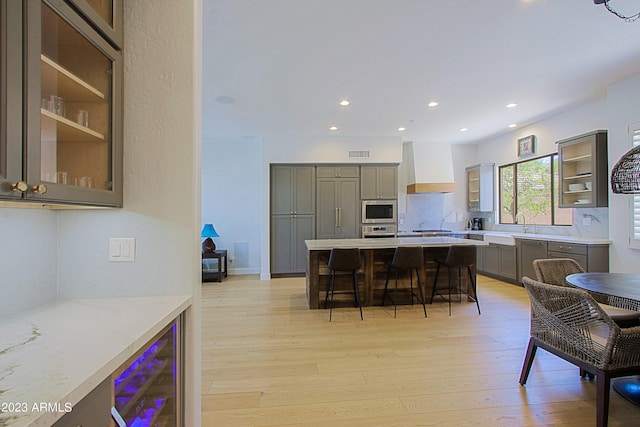 kitchen with appliances with stainless steel finishes, a breakfast bar area, custom exhaust hood, wine cooler, and light hardwood / wood-style flooring