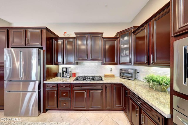 kitchen featuring light stone countertops, stainless steel appliances, decorative backsplash, light tile patterned floors, and glass insert cabinets