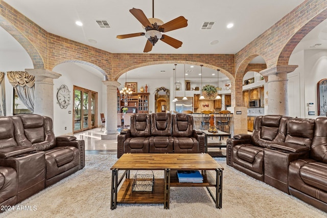 carpeted living room featuring ceiling fan with notable chandelier, ornate columns, and brick wall