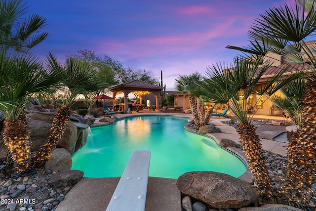 pool at dusk featuring a patio area, pool water feature, a gazebo, and a diving board