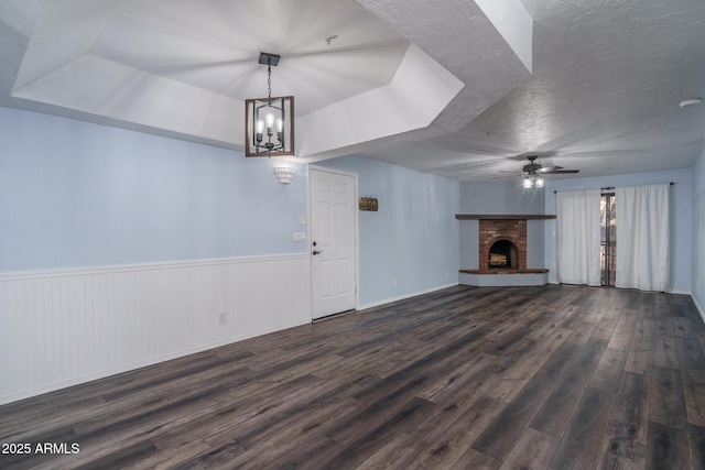 unfurnished living room featuring ceiling fan with notable chandelier, a tray ceiling, a brick fireplace, and dark hardwood / wood-style floors