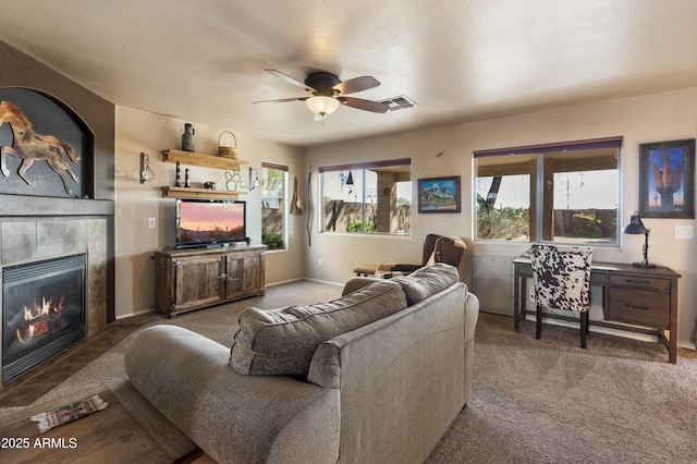 living room with a textured ceiling, ceiling fan, a tiled fireplace, and dark colored carpet