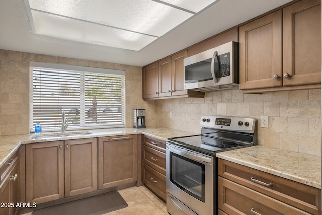 kitchen with brown cabinetry, appliances with stainless steel finishes, light tile patterned flooring, a sink, and backsplash