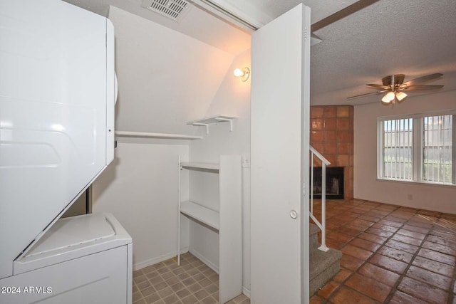 clothes washing area featuring ceiling fan, a textured ceiling, stacked washer / dryer, and a tile fireplace