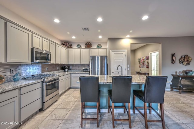 kitchen with gray cabinetry, light stone counters, a kitchen breakfast bar, an island with sink, and stainless steel appliances