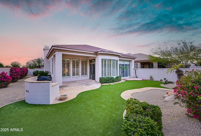 back of house at dusk featuring a tiled roof, fence private yard, stucco siding, a yard, and a patio area