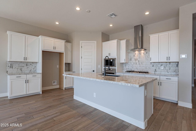 kitchen with white cabinets, stainless steel gas cooktop, light stone counters, an island with sink, and wall chimney range hood