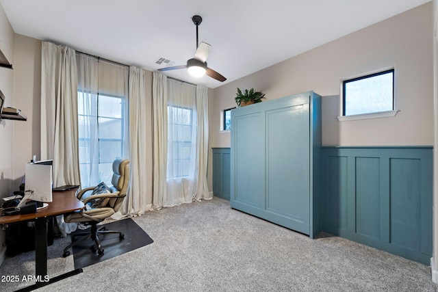 carpeted home office featuring ceiling fan, a decorative wall, visible vents, and wainscoting