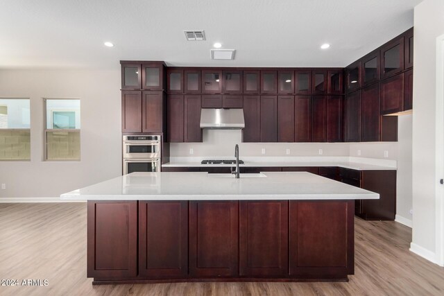 kitchen with light wood-type flooring, double oven, sink, exhaust hood, and a center island with sink