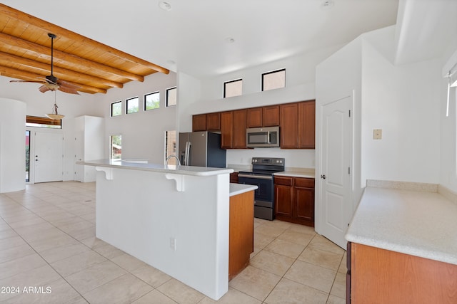 kitchen with stainless steel appliances, an island with sink, beamed ceiling, light tile patterned floors, and ceiling fan